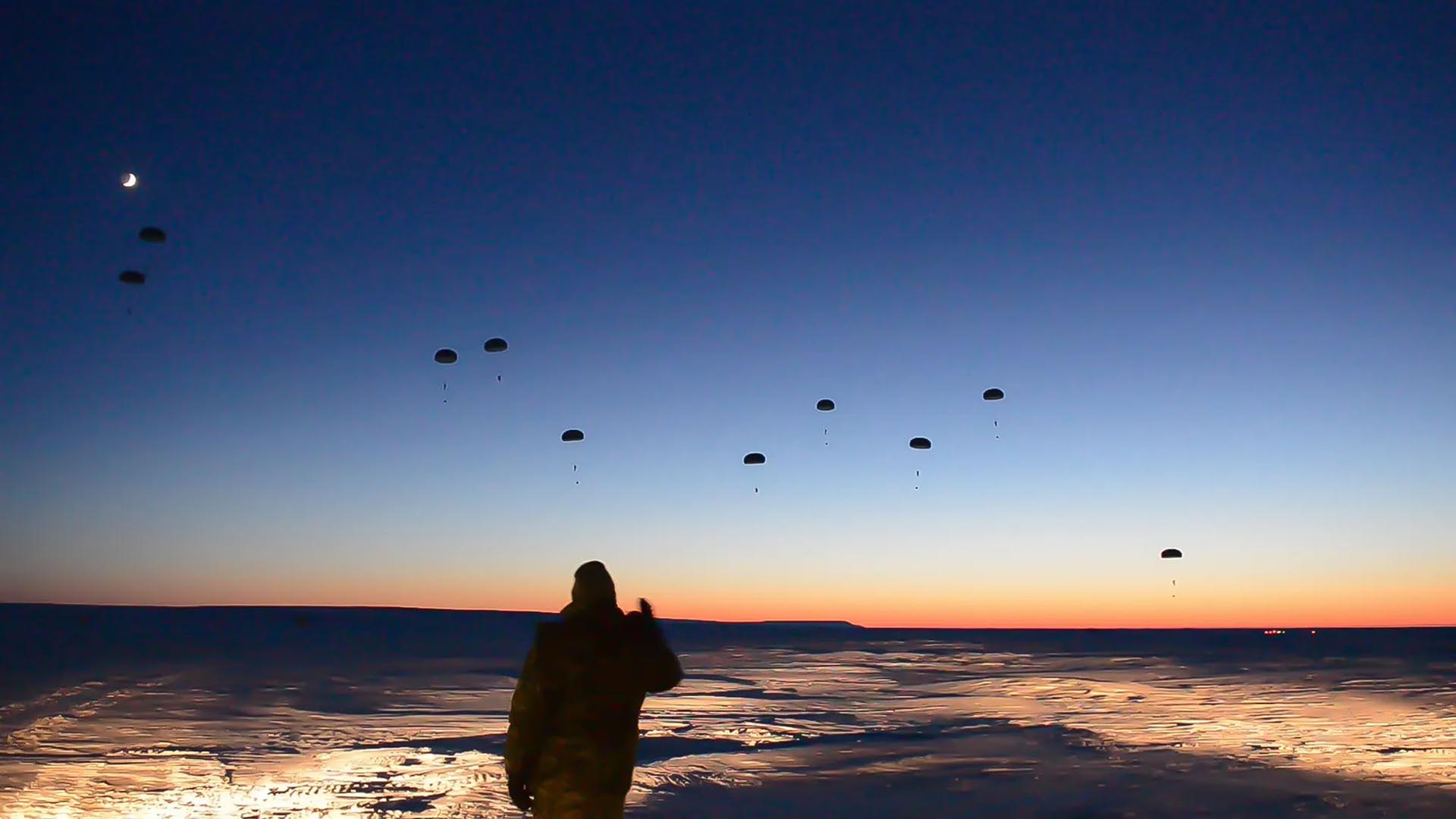 A figure watches as soldiers parachute down to the ground as part of military exercise